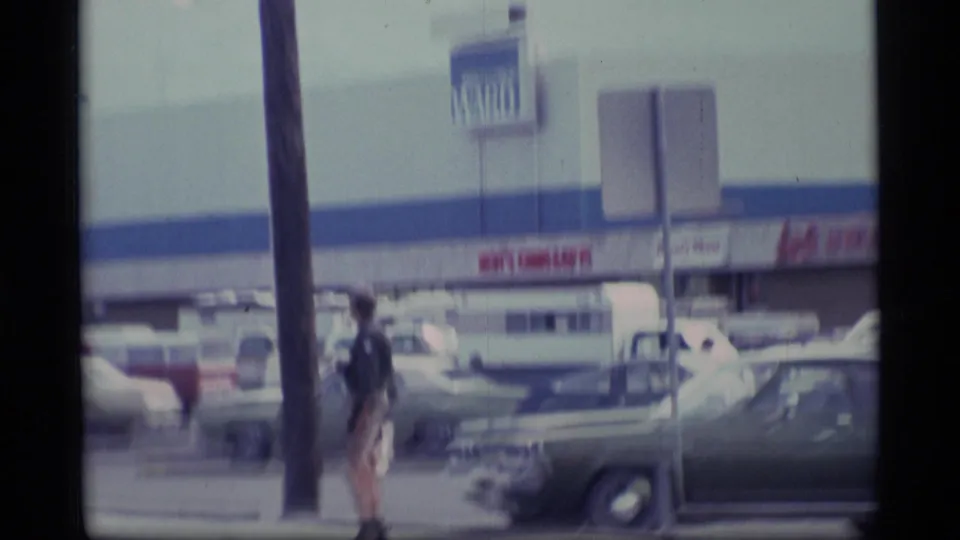 Soft 8mm frame of a 1977 Alaska parking lot and storefront, with cars and a woman walking along the sidewalk.