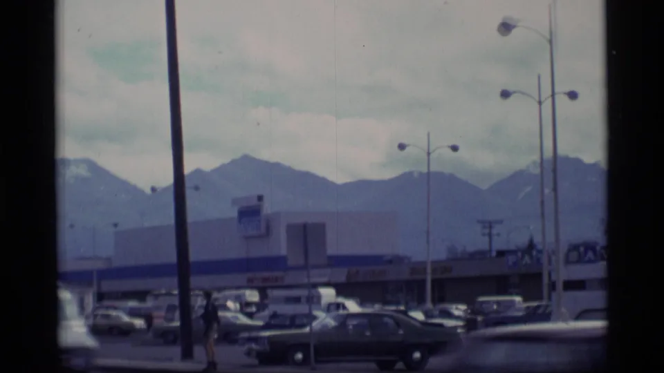 1977 Alaska street-side view of shops, parked cars, tall lamps, signs, and distant mountains under cloudy sky.