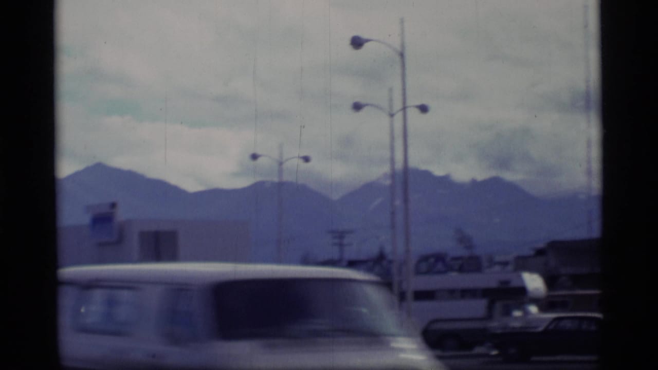 1977 Alaska home-movie frame showing a roadside area with parked vehicles, streetlights, low buildings, and a mountain backdrop.