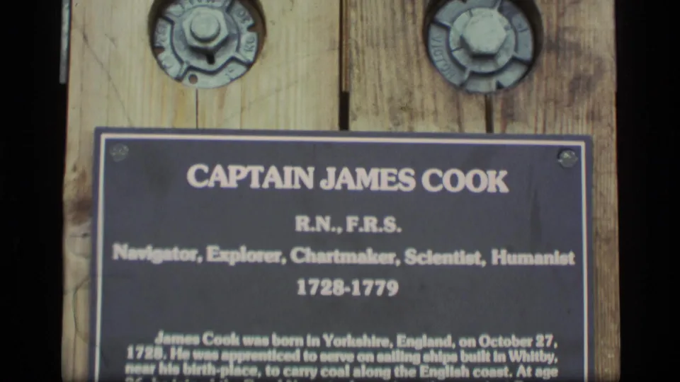 Close view of a Captain James Cook commemorative plaque on weathered wood, with large round bolts visible above the marker.