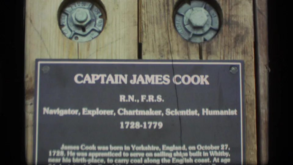 Close 8mm frame of the Captain James Cook commemorative plaque in Alaska, showing his listed roles and lifespan on a wooden mount.