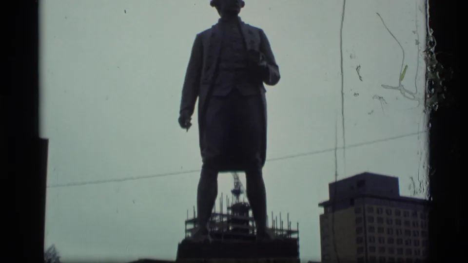 1977 Alaska 8mm frame showing the Captain James Cook statue silhouetted against an overcast sky with urban buildings behind it.
