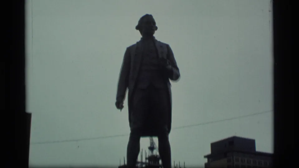1977 Alaska 8mm frame with a low-angle visitor view of the Captain James Cook statue, pale sky, dark frame border, and a building at lower right.
