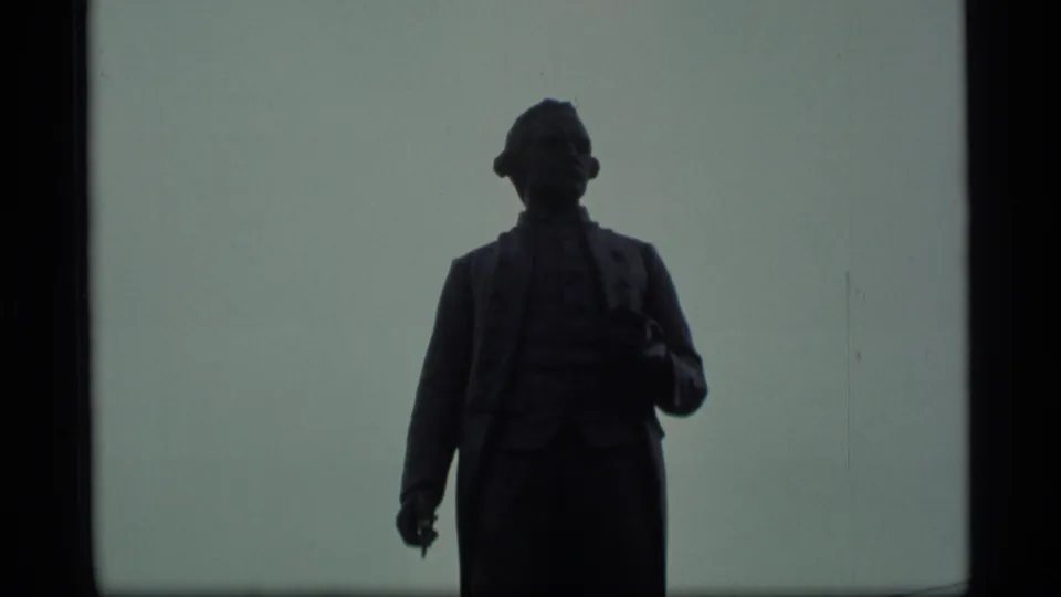 1977 Alaska 8mm frame showing the upper body of the Captain James Cook statue in silhouette against a muted overcast sky.