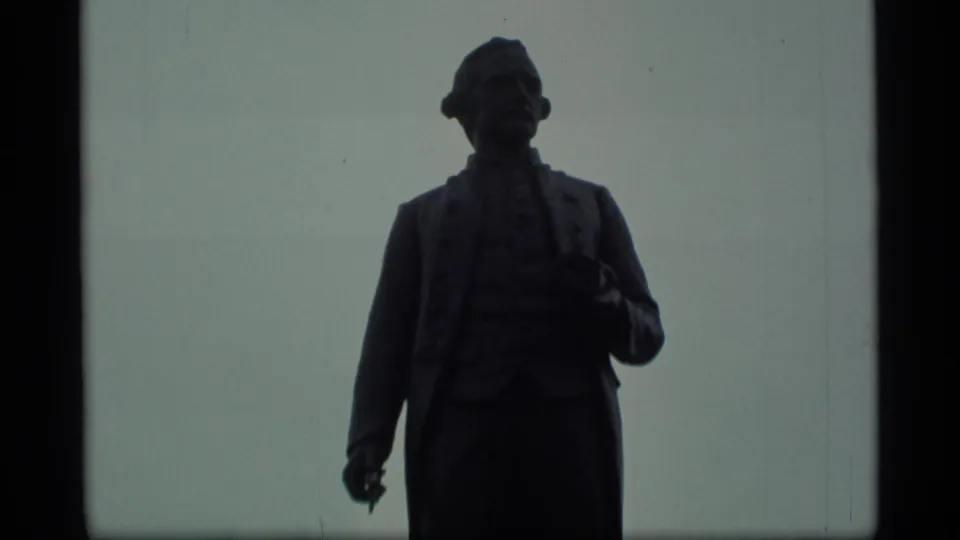 Silhouetted statue in 1977 Alaska home-movie footage, framed close against a grey sky.