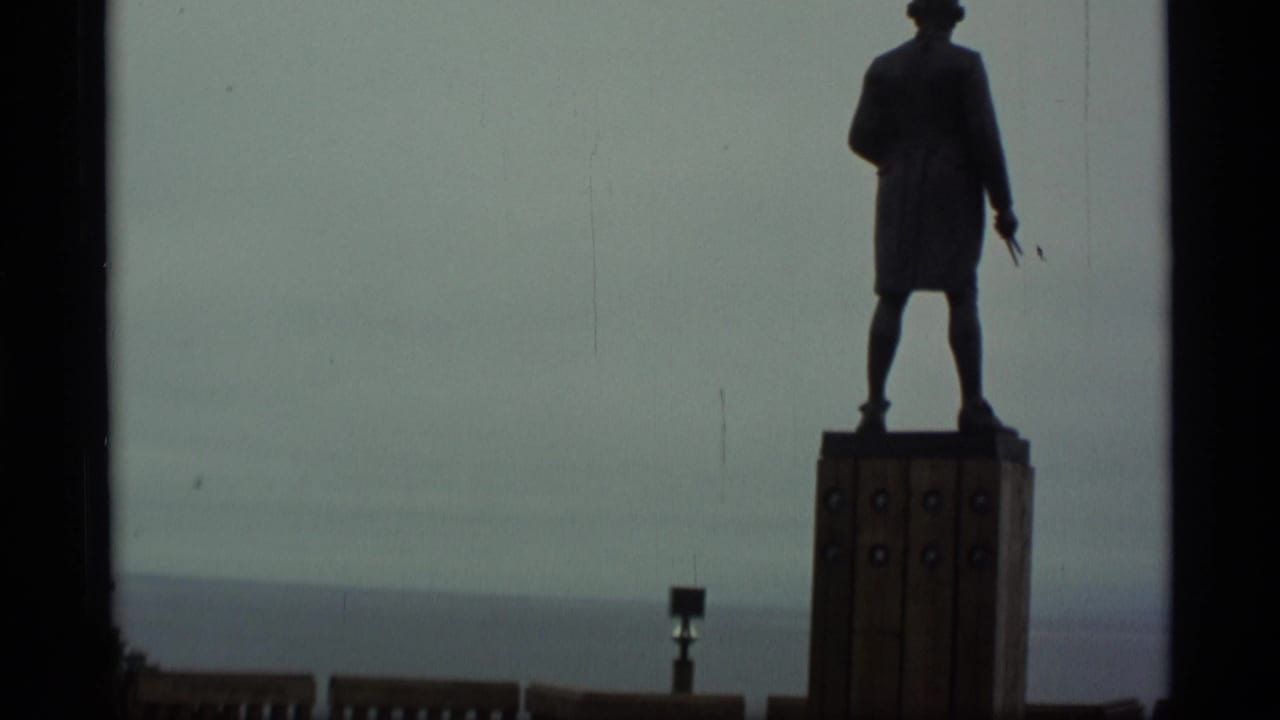 1977 Alaska 8mm frame showing a dark statue on a wood-faced pedestal above a waterfront horizon.