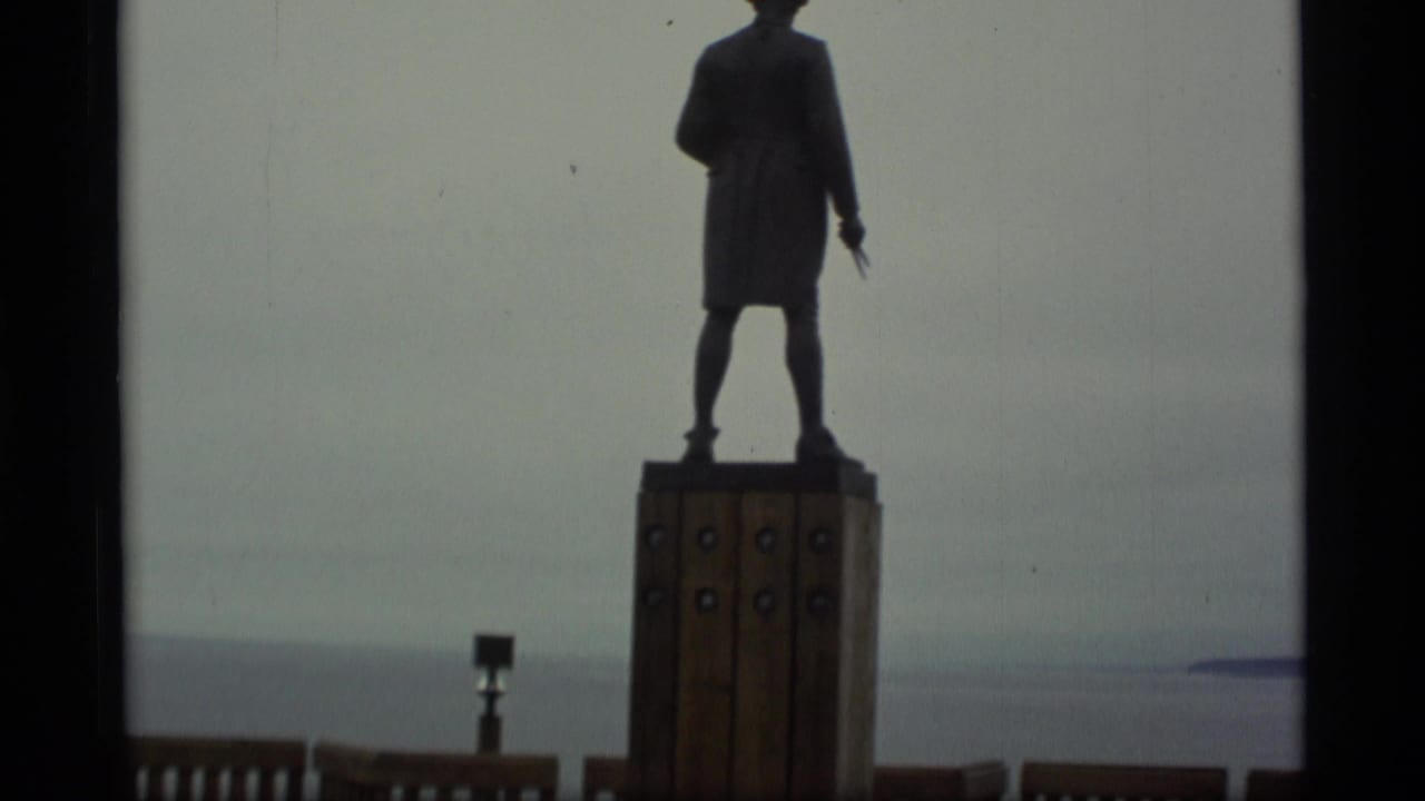 1977 Alaska home-movie still of a silhouetted figure statue facing a broad body of water.