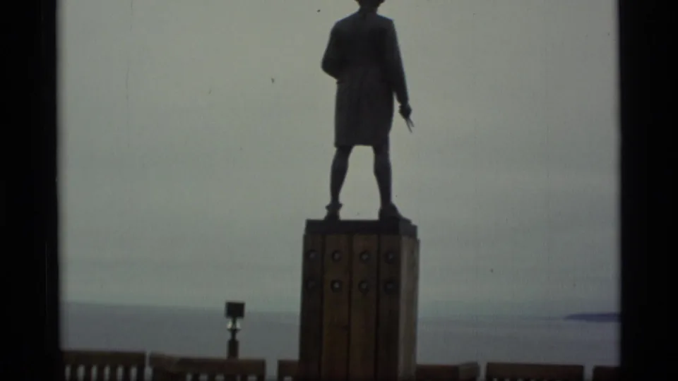 1977 Alaska home-movie still of a silhouetted figure statue facing a broad body of water.