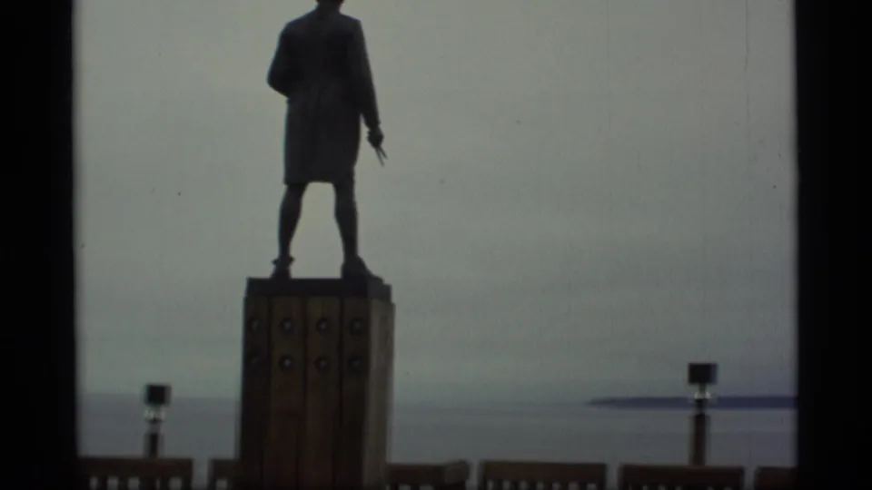 1977 Alaska 8mm frame with the statue partly cropped above a tall pedestal beside an open-water view.