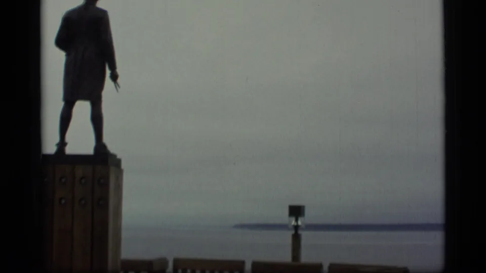 1977 Alaska home-movie frame showing the waterfront statue shifted left as the camera pans across the horizon.
