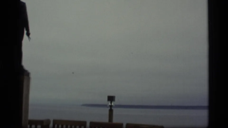 1977 Alaska 8mm frame looking across gray water from a wooden overlook, with distant shoreline and dark film-edge framing.