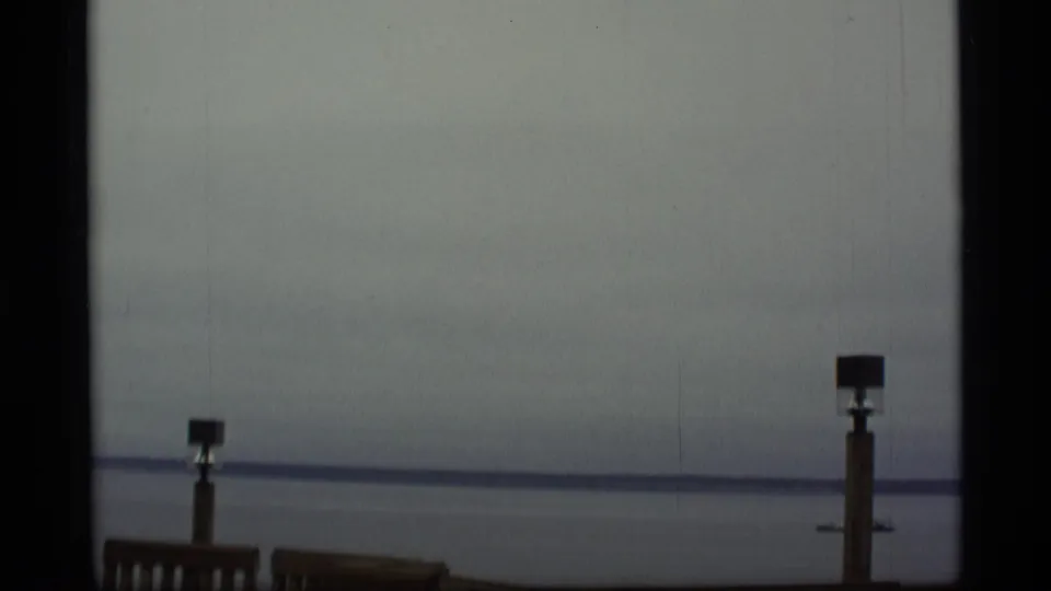 1977 Alaska 8mm frame with a hazy ocean horizon, simple railing, and a post silhouetted against muted water.