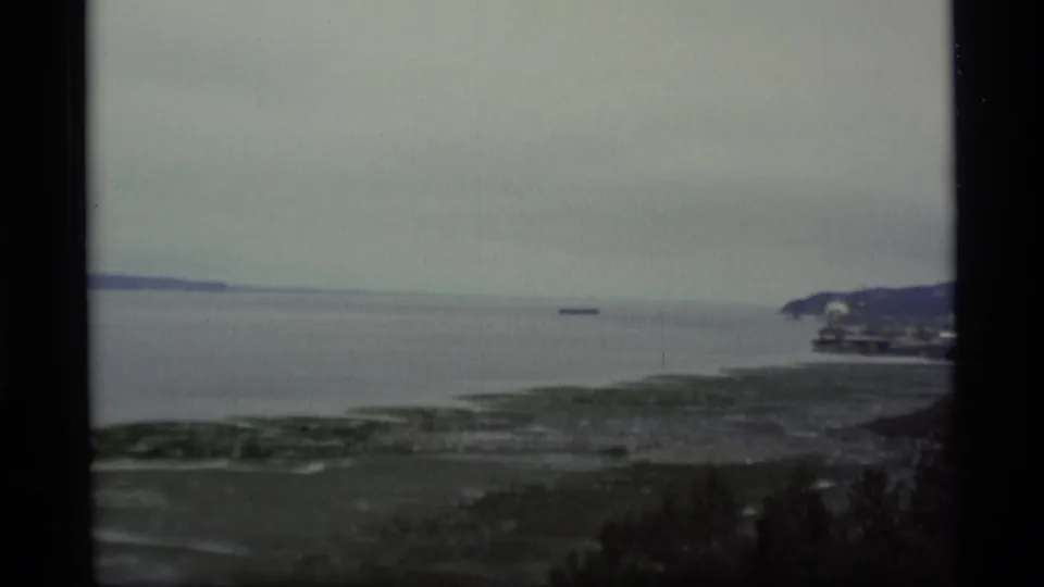 1977 Alaska 8mm view across a calm overcast bay toward a distant vessel, shoreline, and low coastal hills.