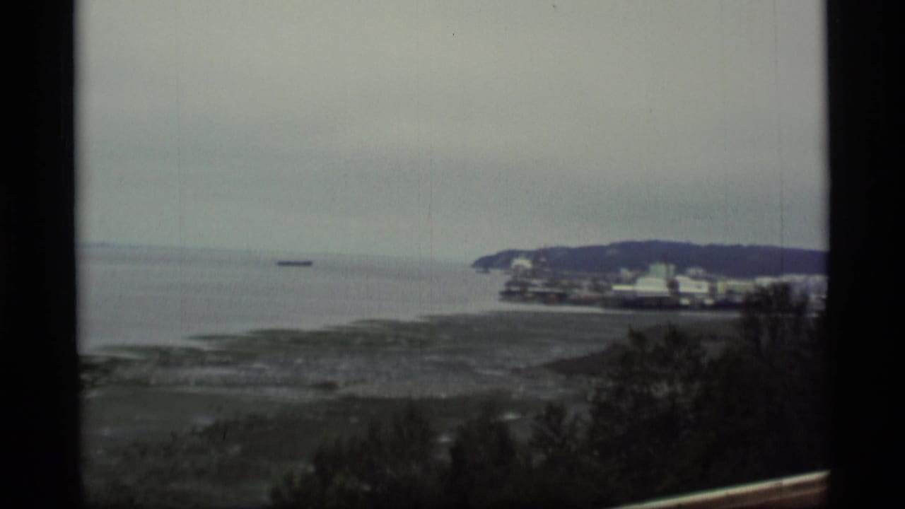 Bay and rocky shoreline with a distant vessel, low hill, and waterfront buildings at right.