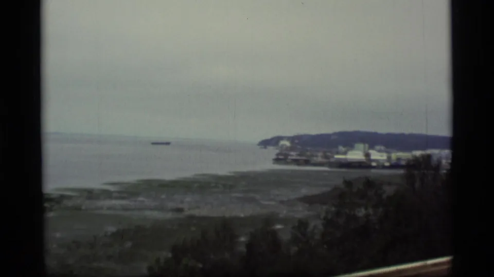 1977 Alaska home-movie frame showing the pan across the bay toward waterfront buildings and a low headland.