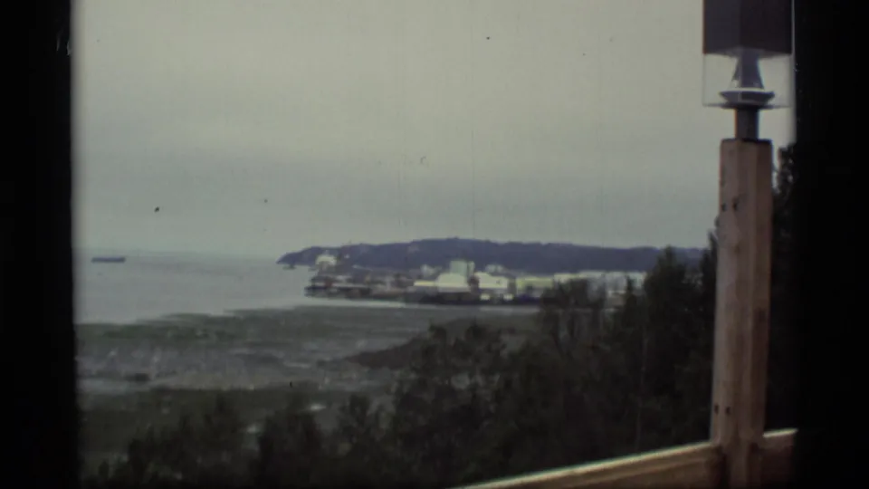 1977 Alaska 8mm frame with the waterfront beyond dark foreground trees, a railing, and a vertical post.