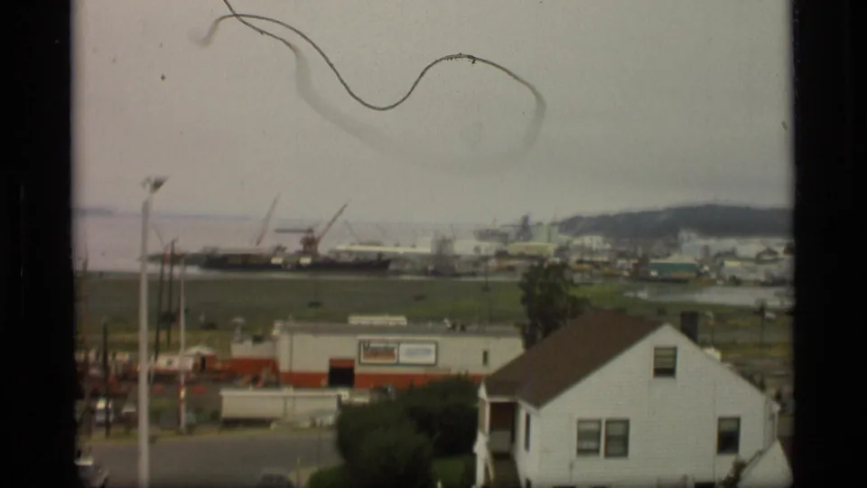 1977 Alaska frame showing a working port with cranes and docked vessels behind roadside buildings and a white house.