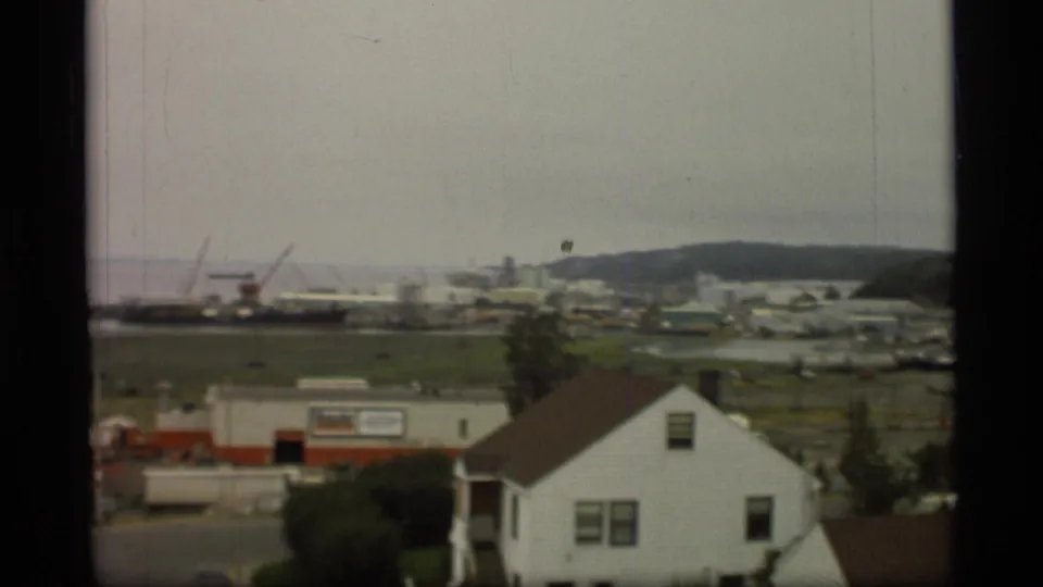 1977 8mm view in Alaska from above white houses toward fields, cranes, ships, and low hills under an overcast sky.