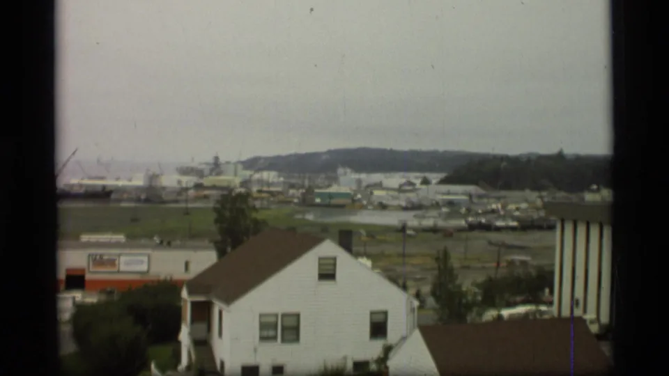 1977 Alaska home-movie frame showing a white house below an elevated viewpoint, with fields, harbor vessels, and hills beyond.
