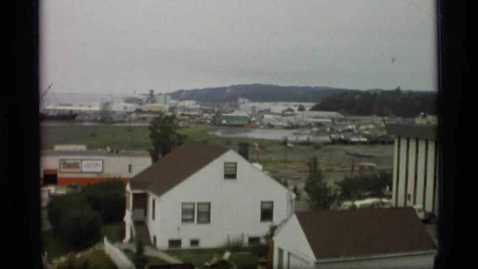 1977 Alaska 8mm frame looking over white houses toward a working harbor, low buildings, cranes, and a muted coastal horizon.