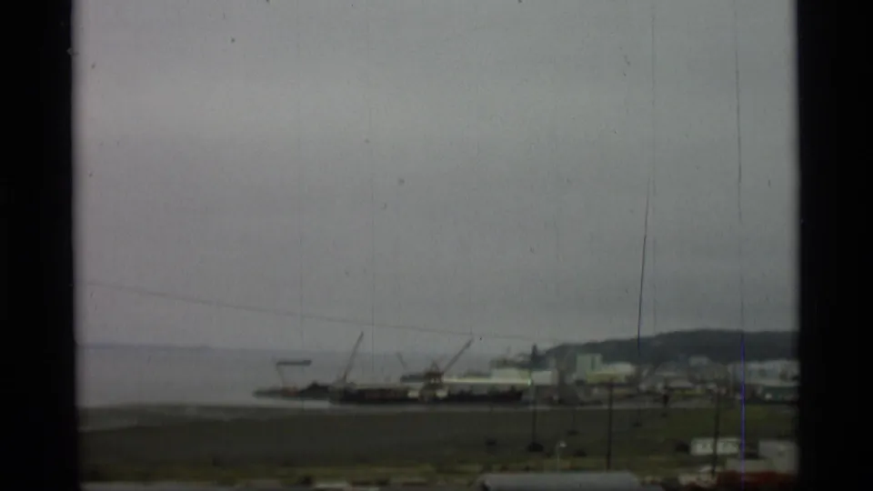 1977 Alaska 8mm frame of a working harbor seen from above, with cranes, boats, shoreline buildings, open grass, and distant hills.