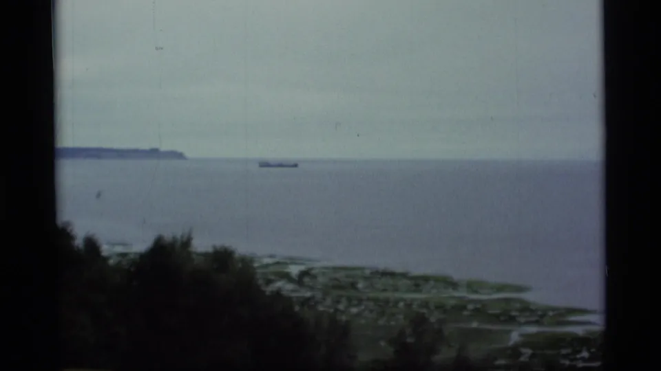 1977 Alaska coastal frame showing calm ocean, a distant vessel, wooded bluff, shoreline, and muted blue-gray overcast light.