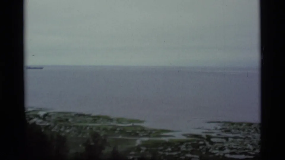 1977 Alaska coastal view with muted gray water, low green shoreline, and dark window or film-frame borders.