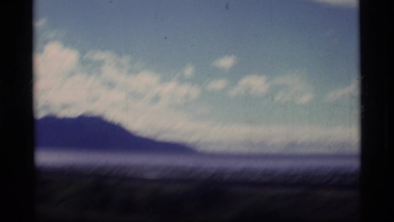 1977 Alaska coastal view with a dark mountain ridge, water, bright blue sky, and scattered white clouds seen through 8mm film softness.
