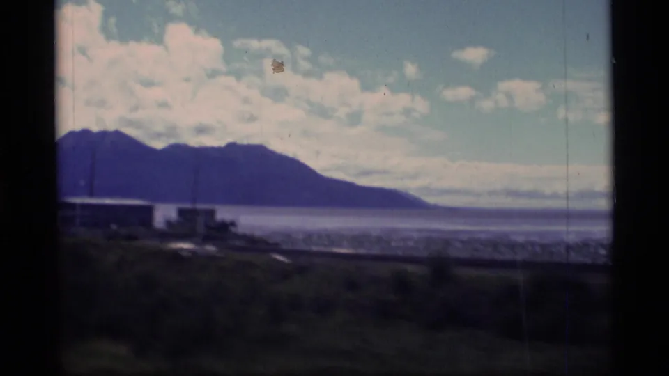1977 Alaska home-movie view of low waterfront buildings, grassy foreground, mountain range, and bright clouded sky along the coast.