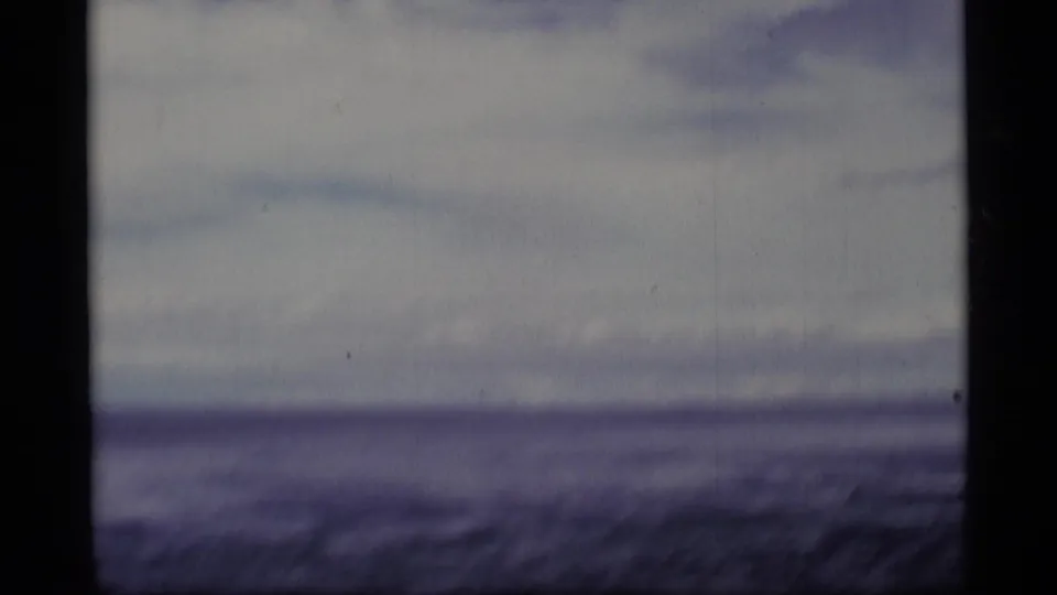 Soft 1977 Alaska coastal frame showing gray water, low horizon, overcast sky, and motion blur from a moving home-movie viewpoint.