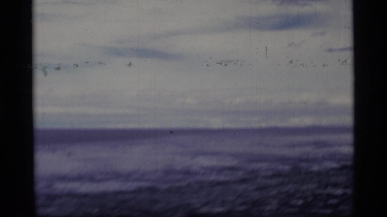 1977 Alaska 8mm frame of a lake and low cloud bands, with grain, scratches, and a dark vertical frame border.