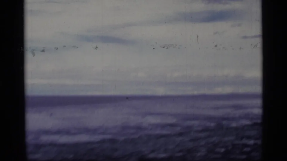 1977 Alaska 8mm frame of a lake and low cloud bands, with grain, scratches, and a dark vertical frame border.