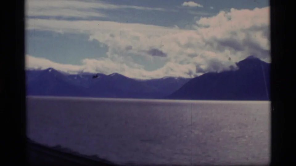 1977 Alaska 8mm frame showing a lake before distant mountain ridges, snow patches, and heavy clouds in a faded home-movie scan.
