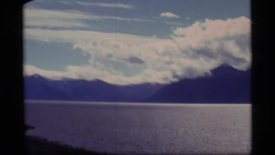 1977 Alaska 8mm frame of a calm lake, dark mountain ridge, and bright cloud cover, with visible grain and faded color.