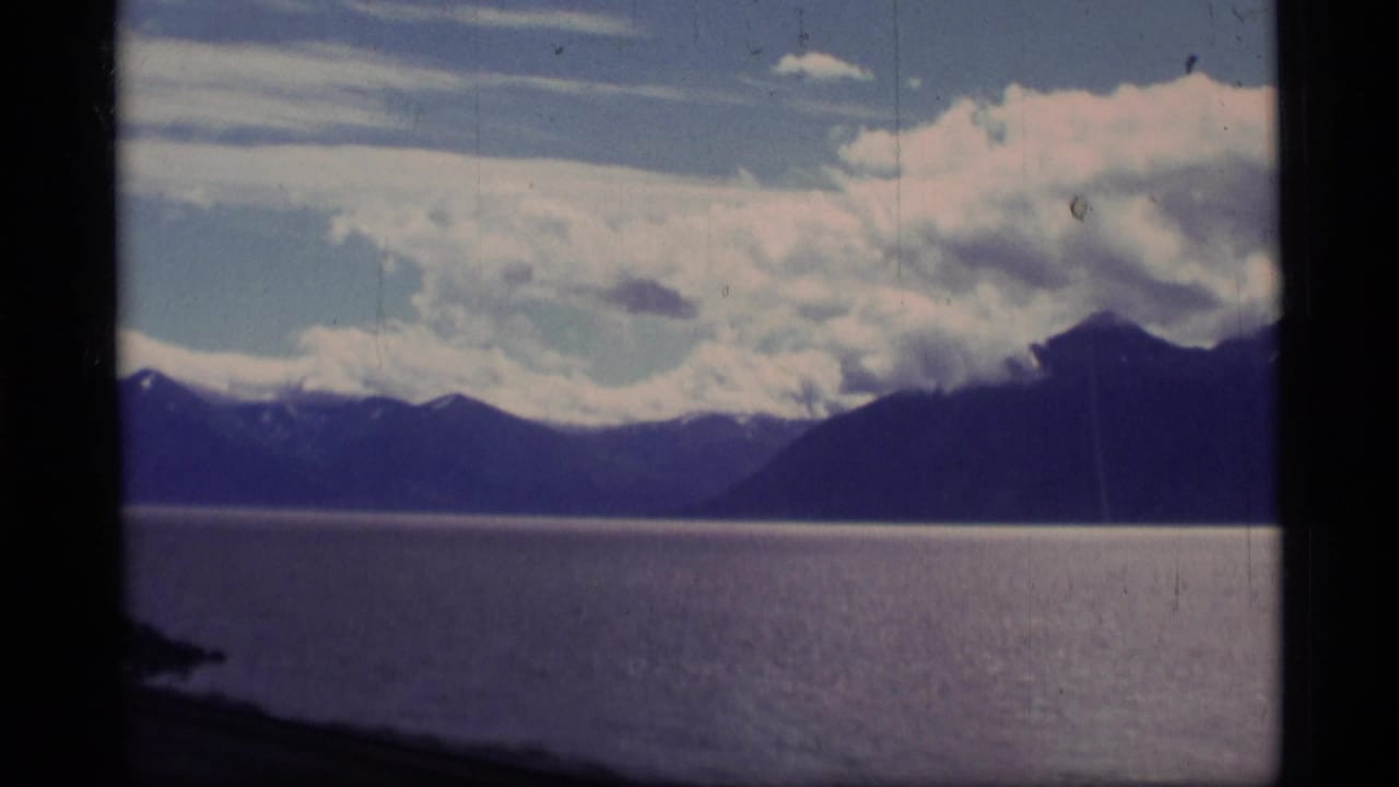 1977 Alaska 8mm frame showing open water, distant mountain ridges, and bright clouds seen from a moving boat.