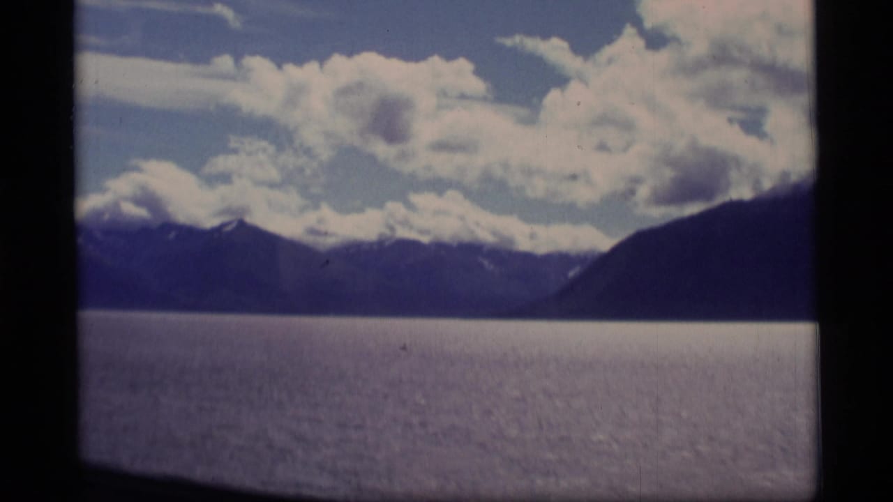 1977 Alaska 8mm boat-view frame with calm water, dark mountain slopes, snow touches, and layered clouds.