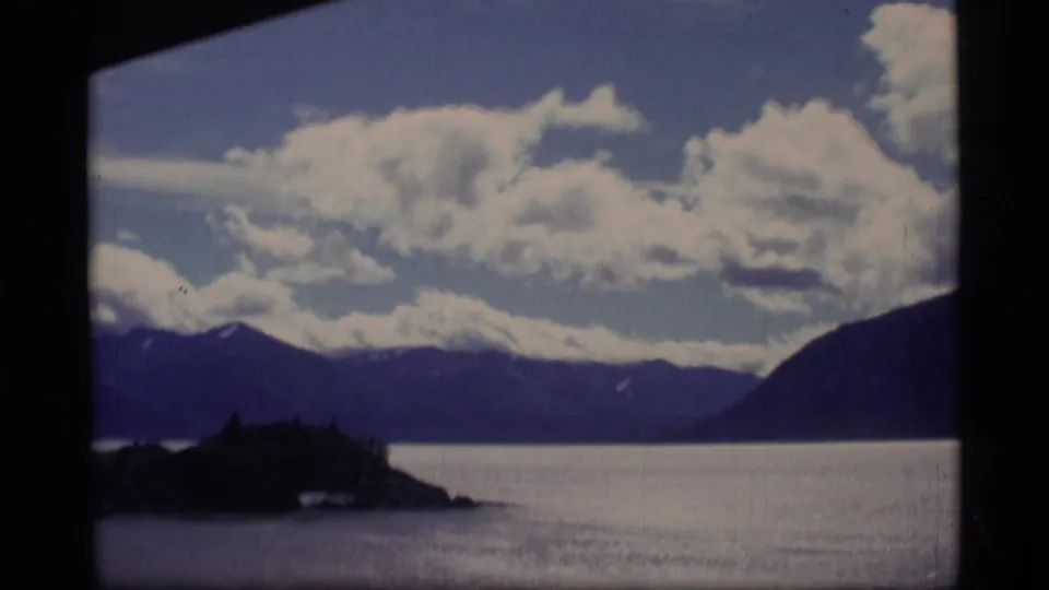 1977 Alaska 8mm boat-view frame with a low forested island, broad water, dark mountains, and bright cloud cover.