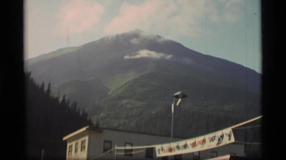 1977 Seward-area view of a green mountain slope, low clouds, street lamp, building, and partial derby banner.