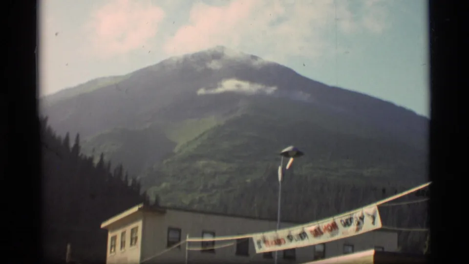 1977 Alaska frame with a clouded green mountain behind a building, street lamp, and partly visible Seward derby banner.