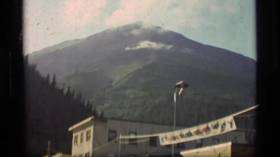 1977 Alaskan home-movie frame showing a green mountain slope behind a low town building, streetlight, and foreground banner.