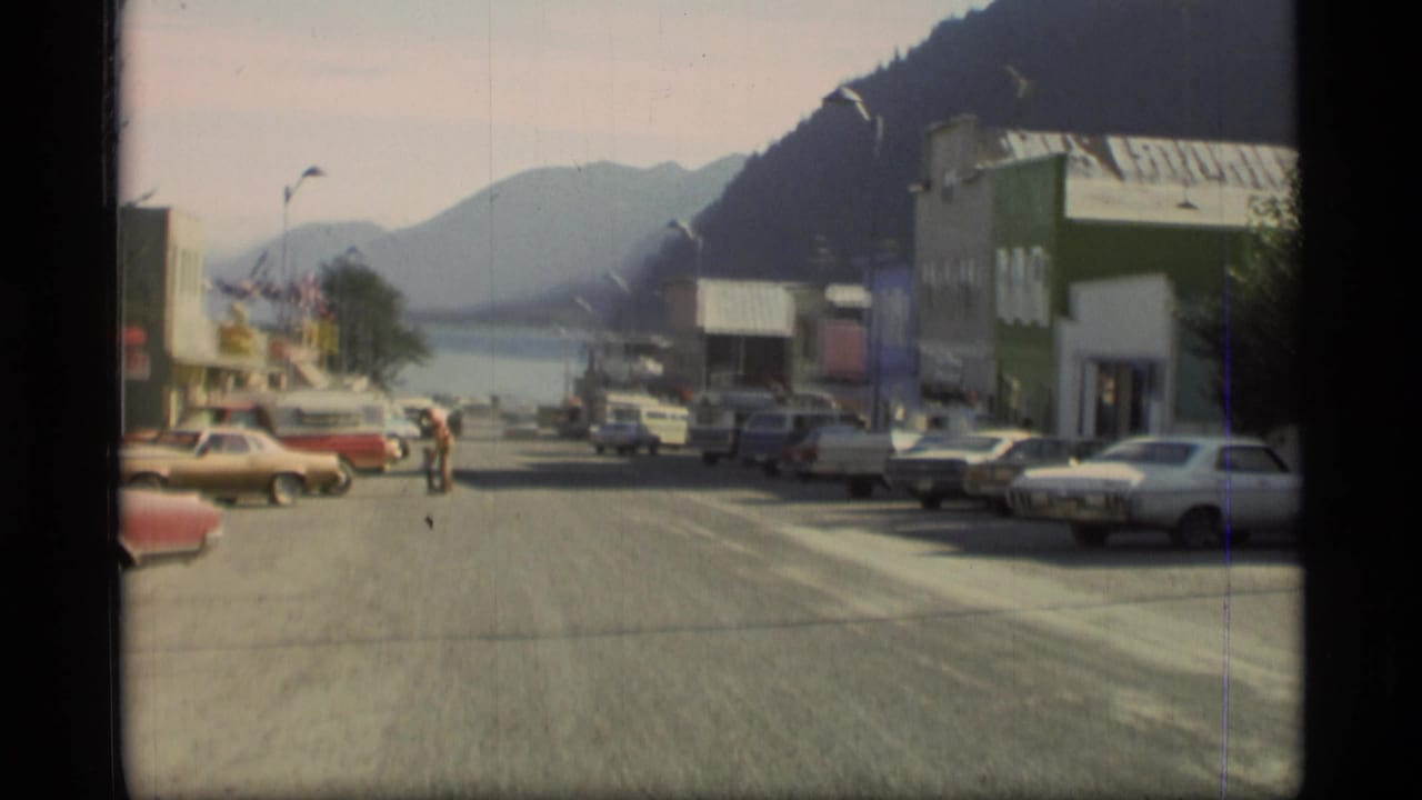 1977 Alaska street view with 1970s cars, storefronts, a person crossing or walking in the road, waterfront, and distant mountains.