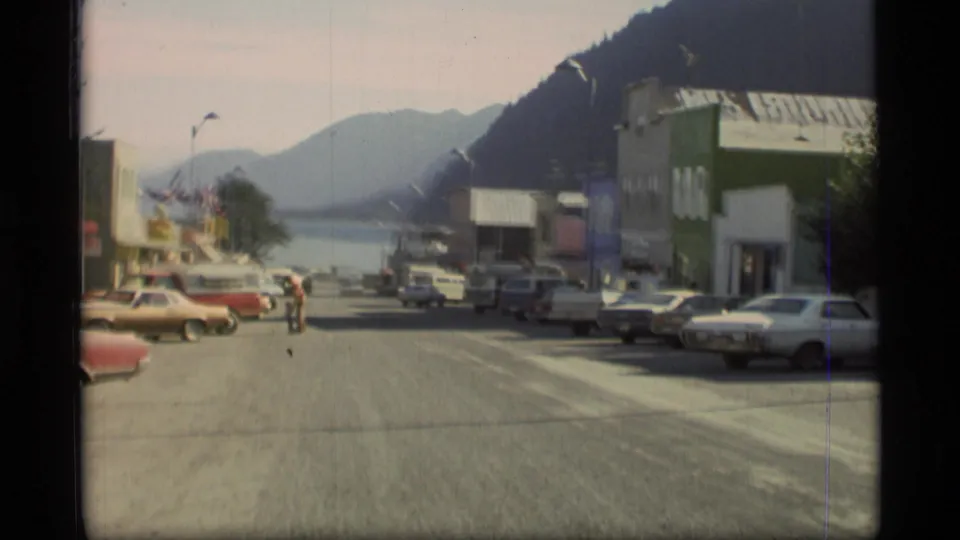 1977 Alaska street view with 1970s cars, storefronts, a person crossing or walking in the road, waterfront, and distant mountains.