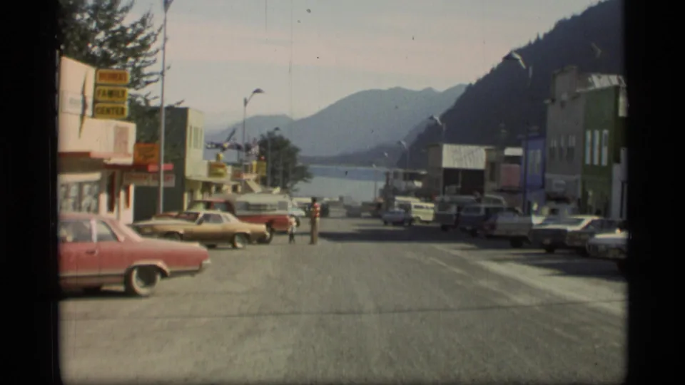 1977 Alaska downtown view with a visible Family Center sign, classic cars, storefronts, a pedestrian, water, and mountain backdrop.