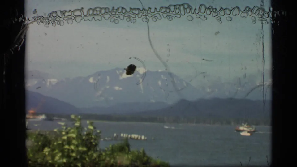 1977 Alaska waterfront frame showing boats on the water, a wooded shoreline, dark hills, and distant snow-marked mountains.