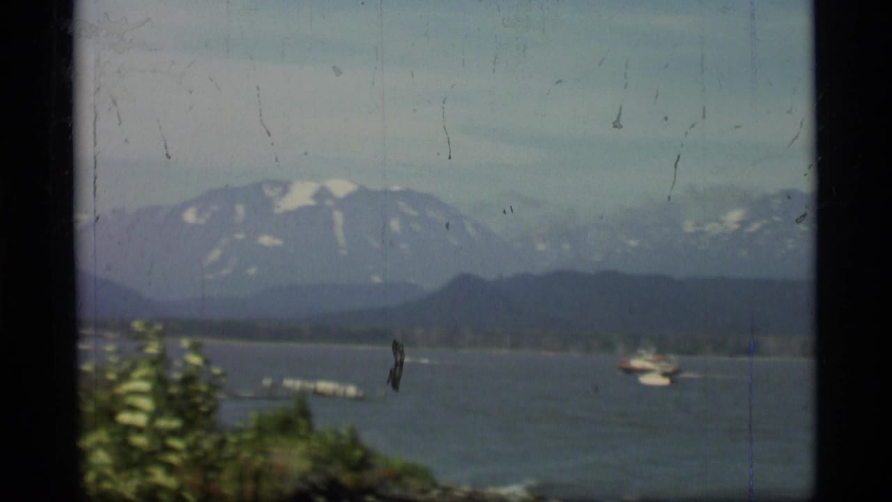 1977 Alaska 8mm frame showing small boats on open water below hazy, snow-streaked mountains.