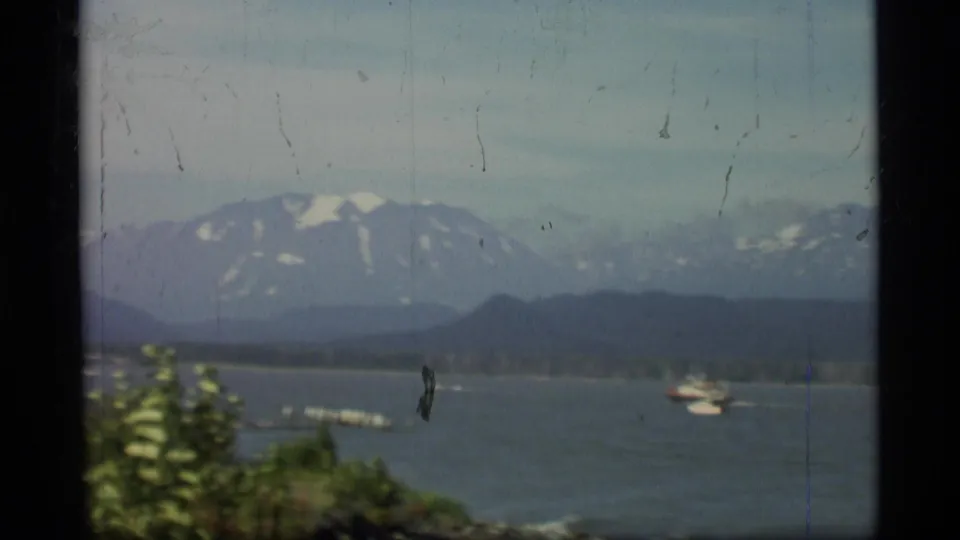 1977 Alaska 8mm frame showing small boats on open water below hazy, snow-streaked mountains.