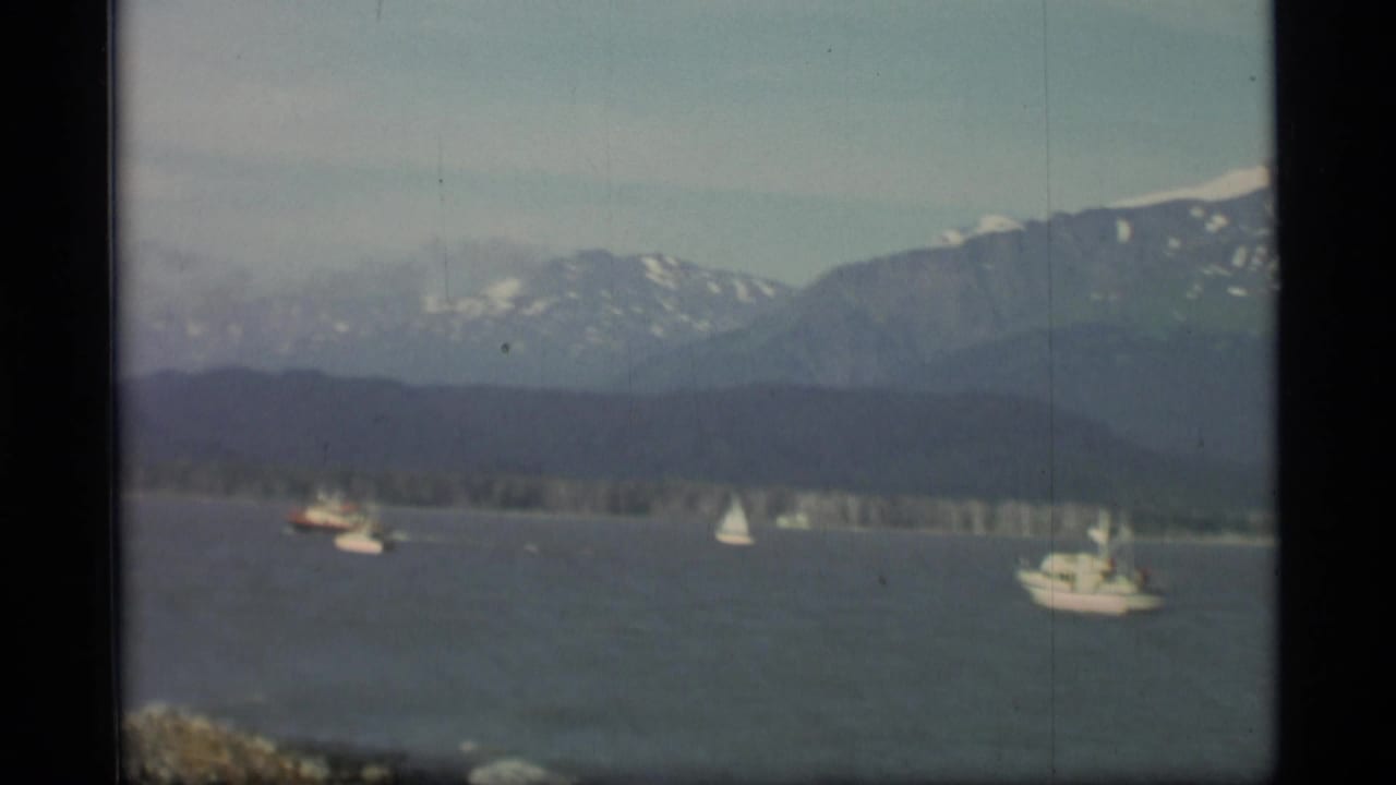 1977 Alaska waterscape with sailboats and a motorboat against a wooded shore and mountain range.