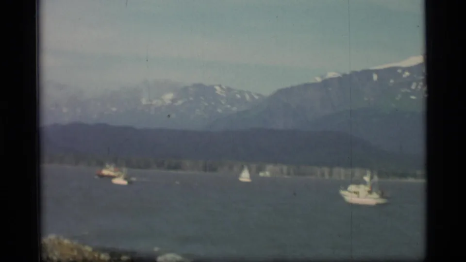 1977 Alaska waterscape with sailboats and a motorboat against a wooded shore and mountain range.
