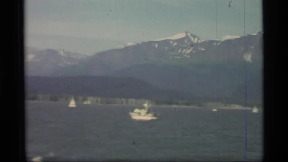 Vintage 8mm Alaska frame with a white motorboat, small sailboats, and snow-marked mountains beyond the shoreline.