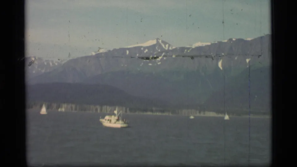 1977 Alaska home-movie frame showing small boats spread across a bay below dark, snow-streaked mountains.